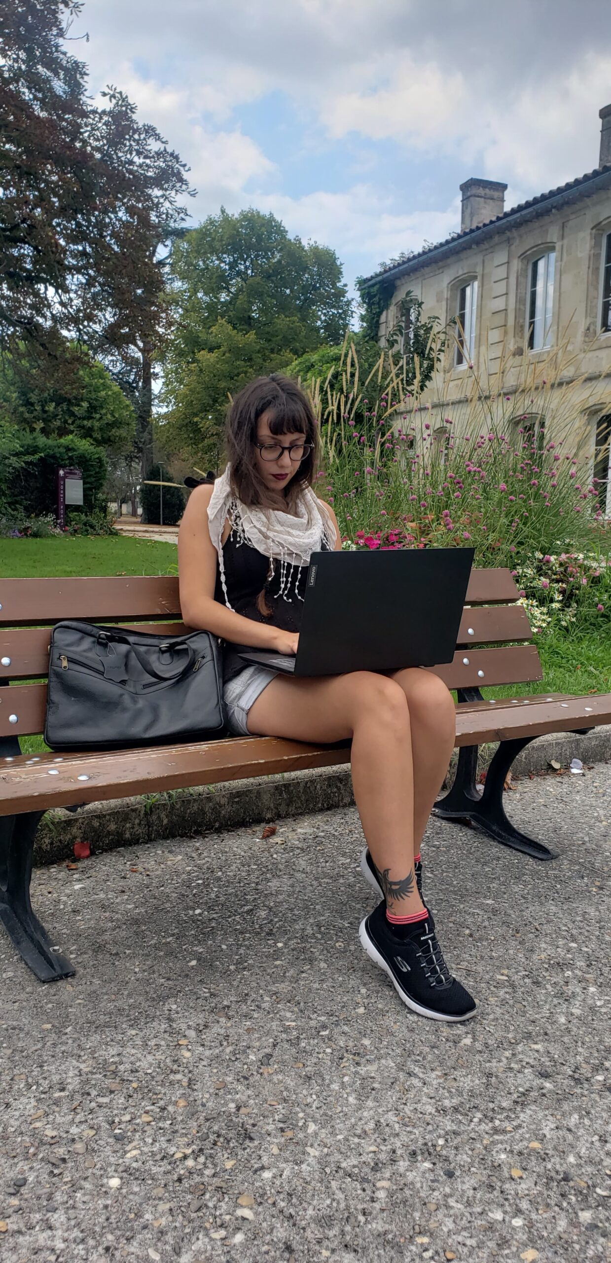 femme sur un banc qui a un pc sur les genoux qui écrit. derrière elle un bâtiment et un parc avec des arbres et des fleurs