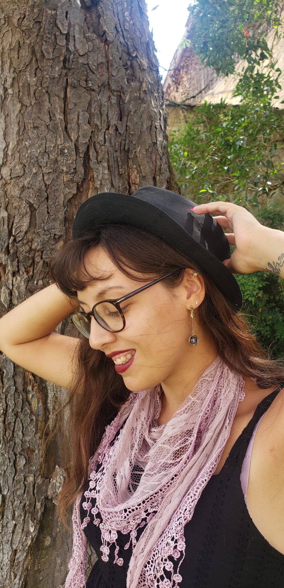 femme de profil qui sourit. elle a des lunettes, un chapeau et un foulard rose. elle est devant un arbre marron .