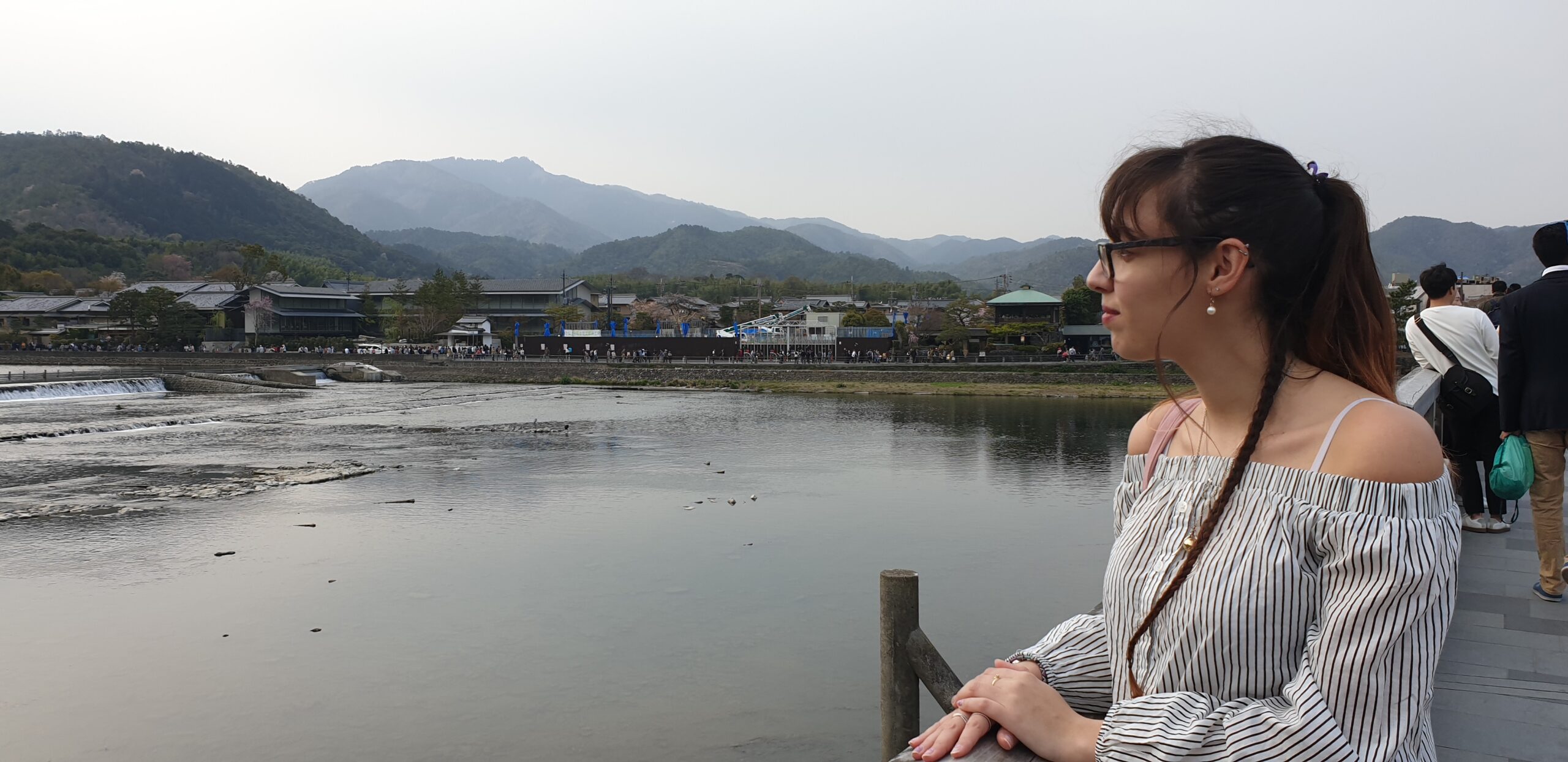 femme de profil cheveux attaché avec une tresse sur le côté elle regarde une rivière calme avec un village et des montagnes en fond.