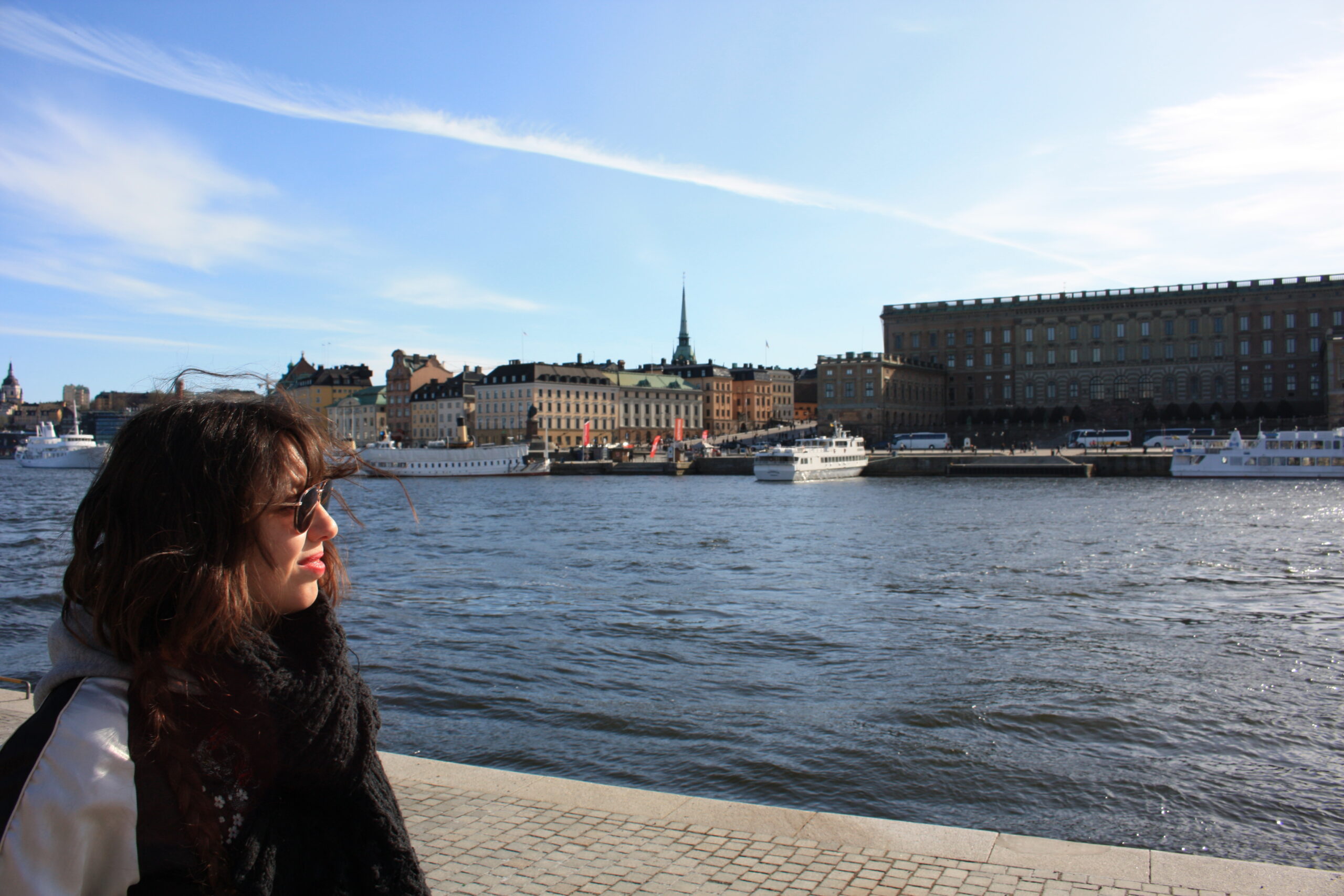 femme de profil avec lunette de soleil noir qui regarde devant elle,. derrière une rivière bleu et derrière des bateau et des bâtiments de couleurs variés : blanc, briques, beiges...