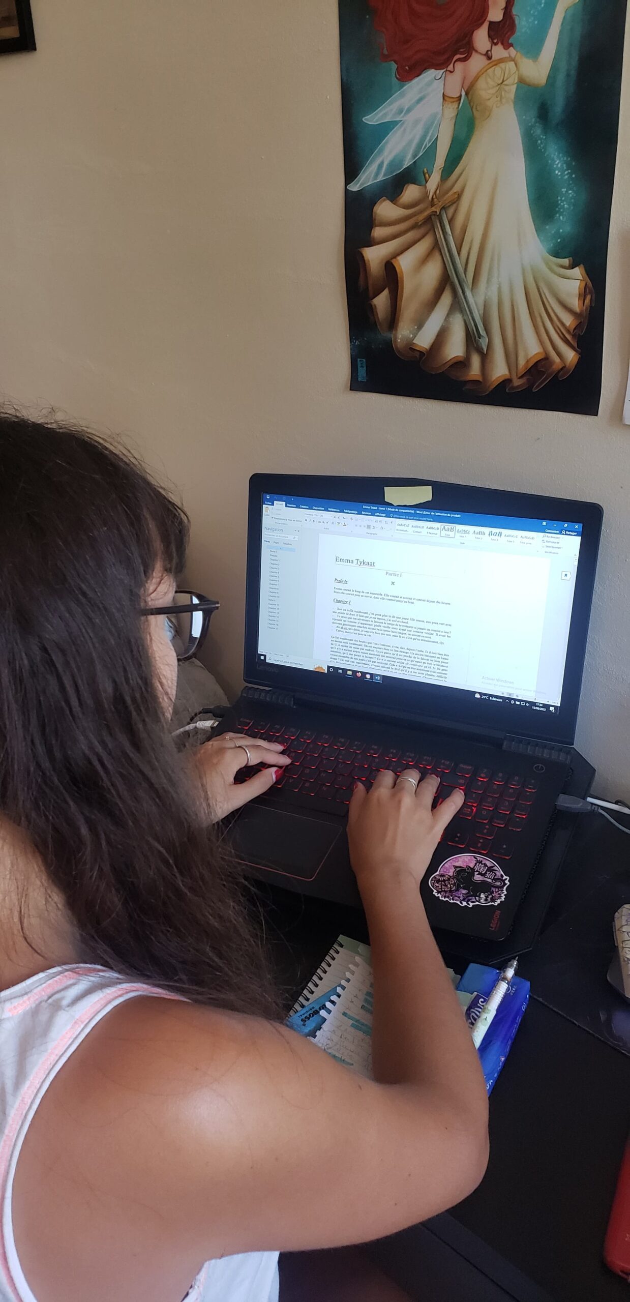 une femme à lunette qui écrit sur un ordinateur avec un poster d'une fée qui tient une épée au dessus de l'ordinateur